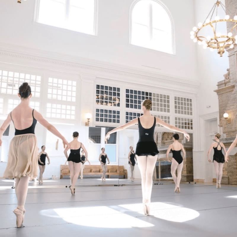 Ballerinas in black leotards practice in a bright, white studio with large windows and a chandelier.