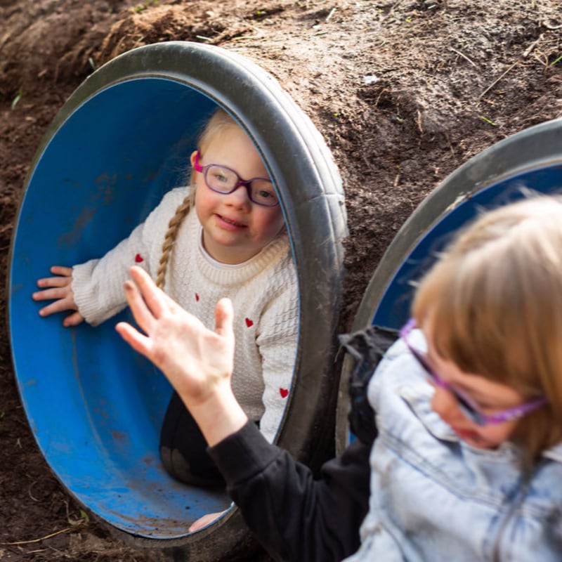 A young girl with glasses smiles from inside a blue plastic tunnel embedded in a dirt hillside, while another child reaches out in the foreground.