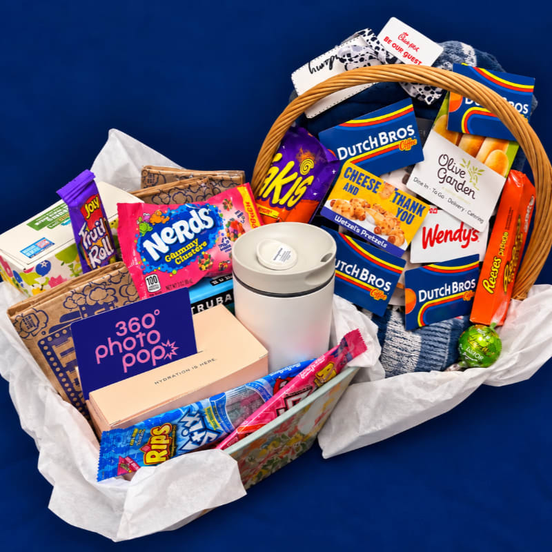 A variety of colorful snacks, gift cards, and a white tumbler are arranged in a white-lined basket against a solid blue background.