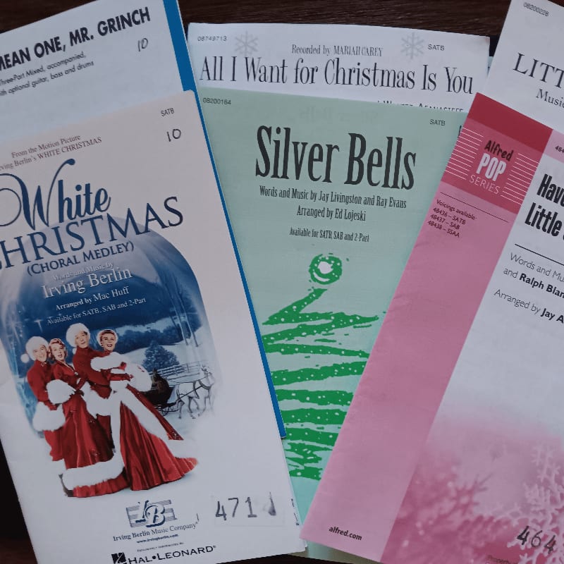 Several sheets of Christmas song music lie scattered across a dark wooden surface.