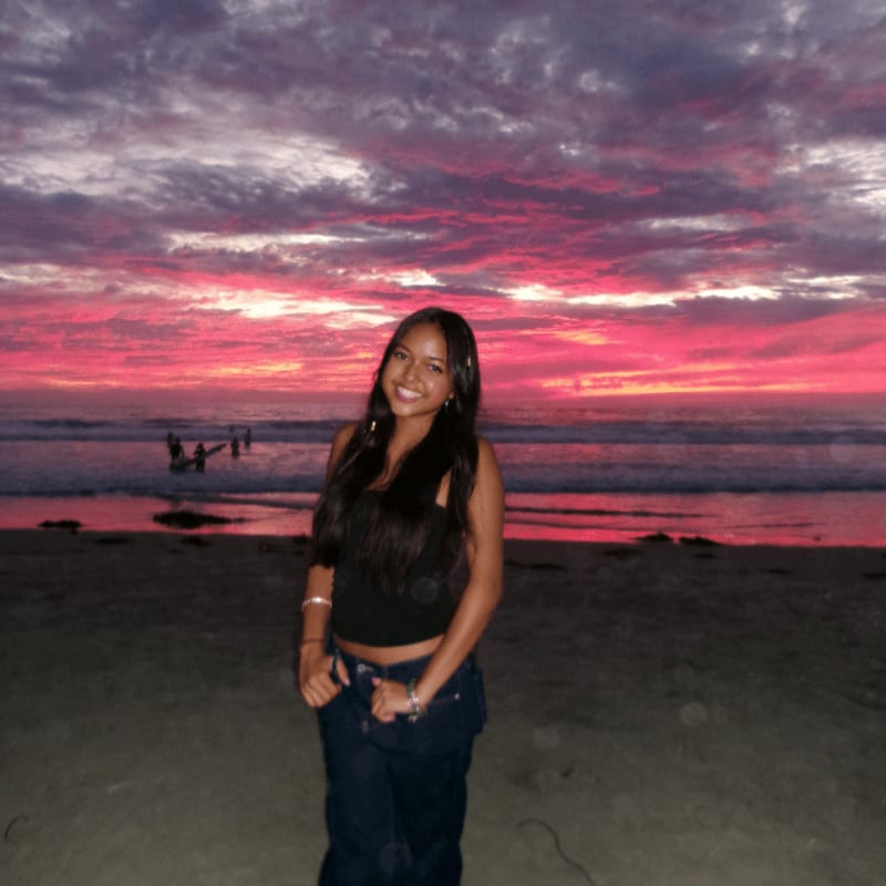 A smiling woman stands on a sandy beach against a backdrop of a vibrant pink and purple sunset over the ocean.