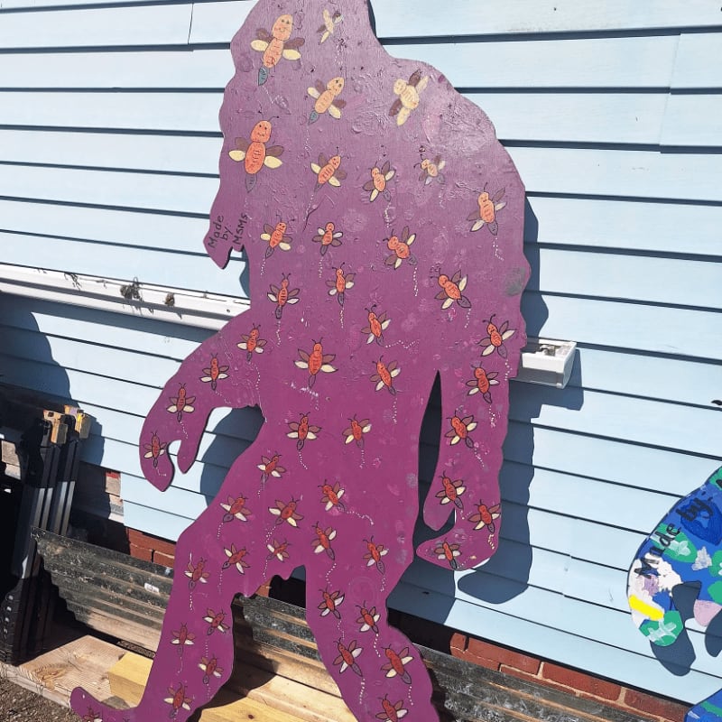 A purple Bigfoot cutout decorated with small bees stands against a light blue wooden siding wall.