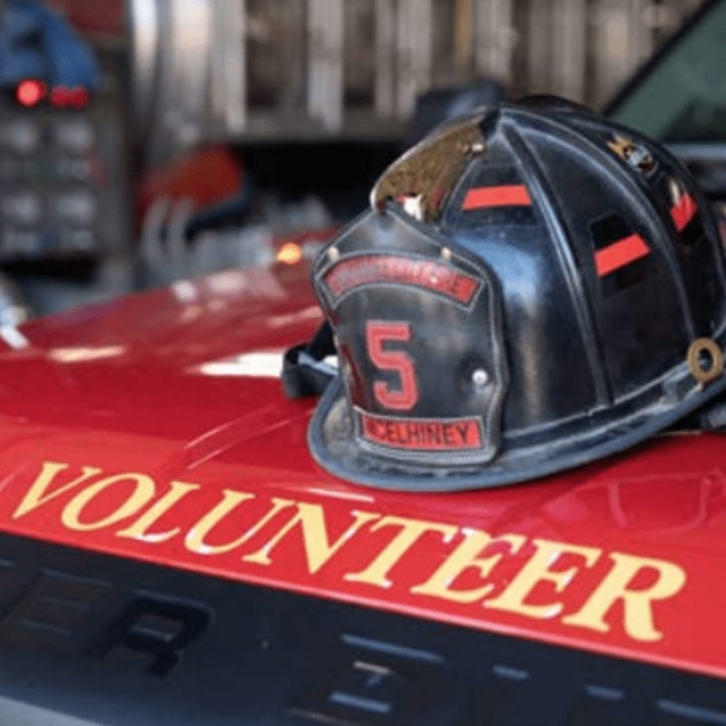 A black firefighter's helmet sits on the red hood of a vehicle labeled "VOLUNTEER" with a blurred fire truck in the background.