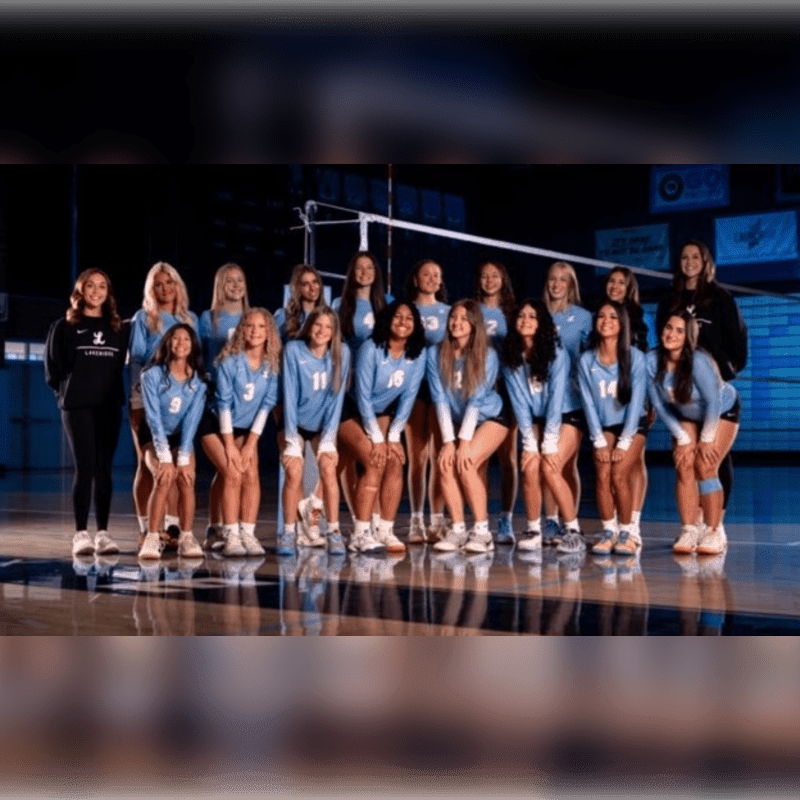 A volleyball team in blue uniforms poses for a group photo on a polished gym floor with a net and dark arena seating in the background.