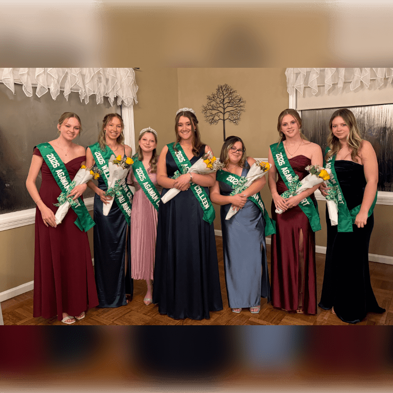 Six young women wearing formal gowns and green sashes stand together in a room with neutral-colored walls and white window valances.