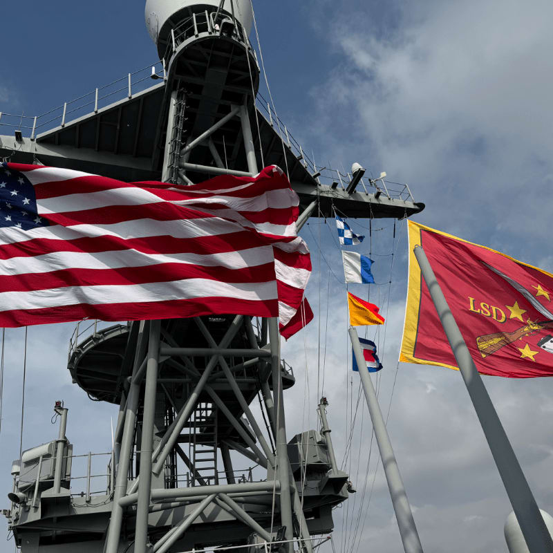 An American flag and a red military flag fly in the foreground against a grey ship structure and a cloudy blue sky.