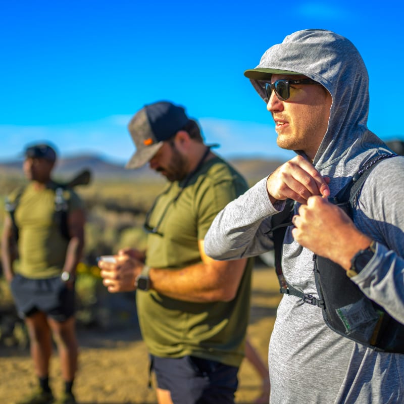 A man in a grey hooded shirt and sunglasses stands in the foreground, with two other men and a desert landscape under a clear blue sky in the background.
