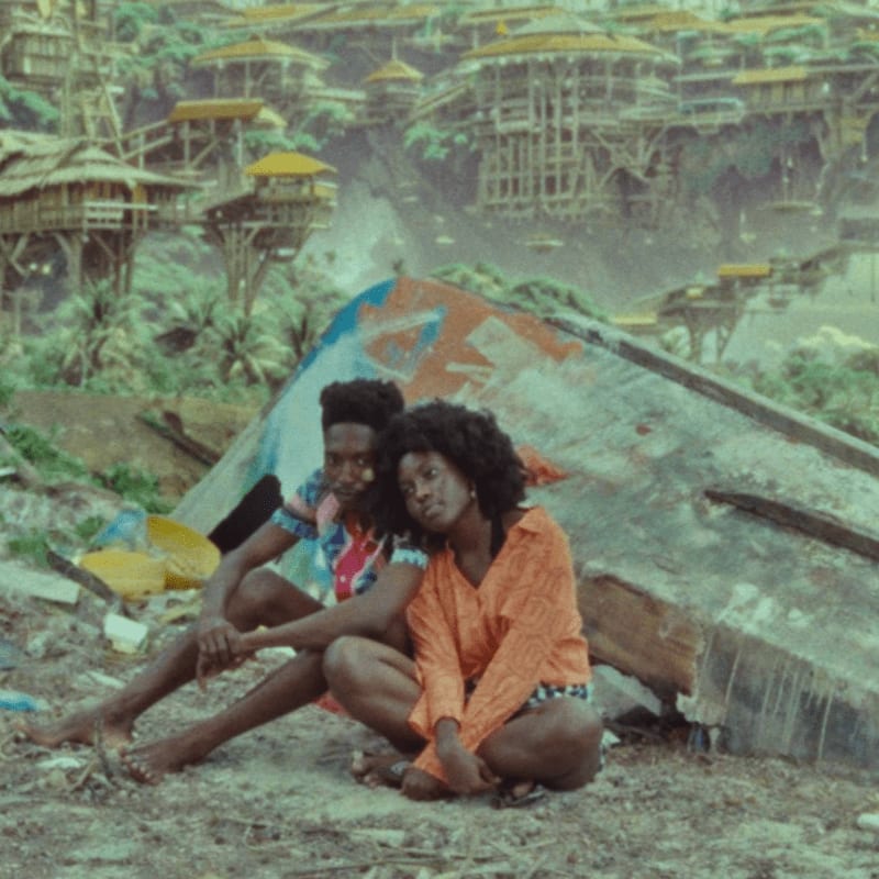 Two people sit on the ground in the foreground, leaning against a weathered structure with a village of stilt houses nestled in a lush green hillside in the background.