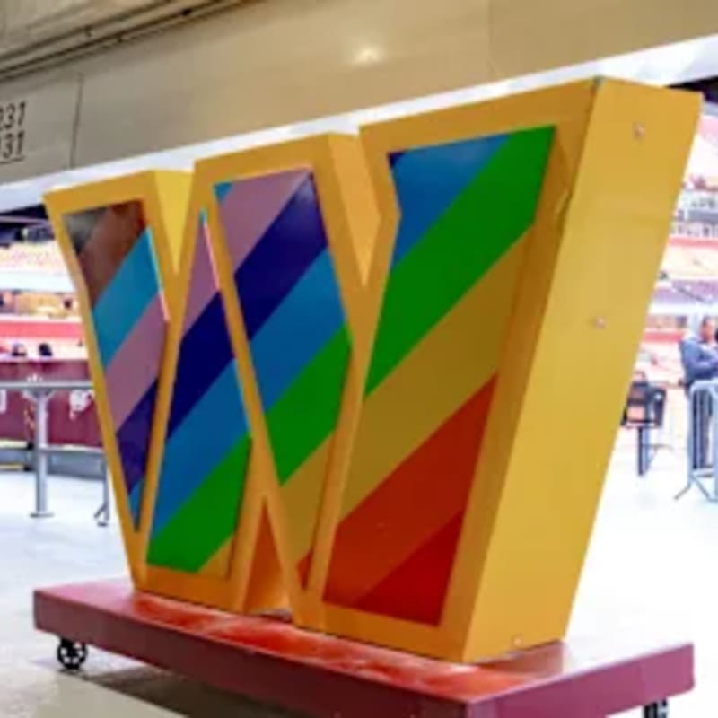 A large, yellow "W" filled with rainbow colors sits on a red wheeled platform in a brightly lit indoor venue.
