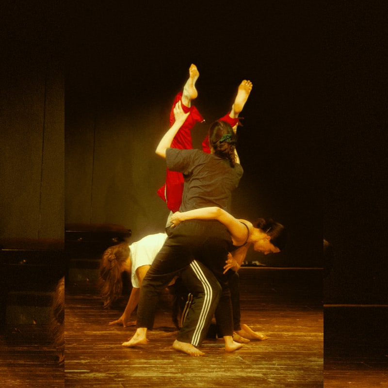 Three dancers perform an acrobatic routine on a wooden stage against a dark, minimalist background.