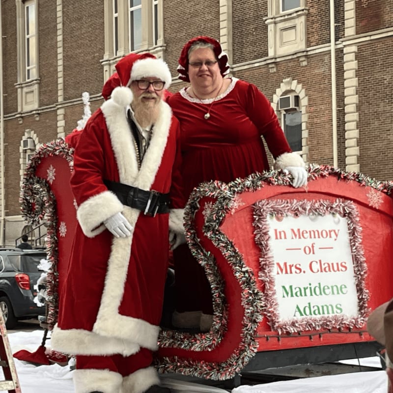 Santa and Mrs. Claus stand beside a red decorative sleigh in front of a brick building.