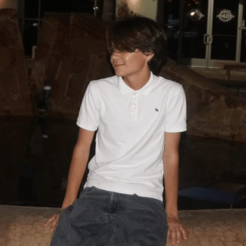 A young man in a white polo shirt and blue jeans sits in the foreground against a background of dark rocks and a glass door.