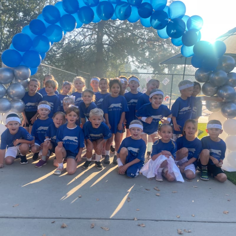 A group of children in blue shirts pose under a blue balloon arch with trees and a fence in the background.