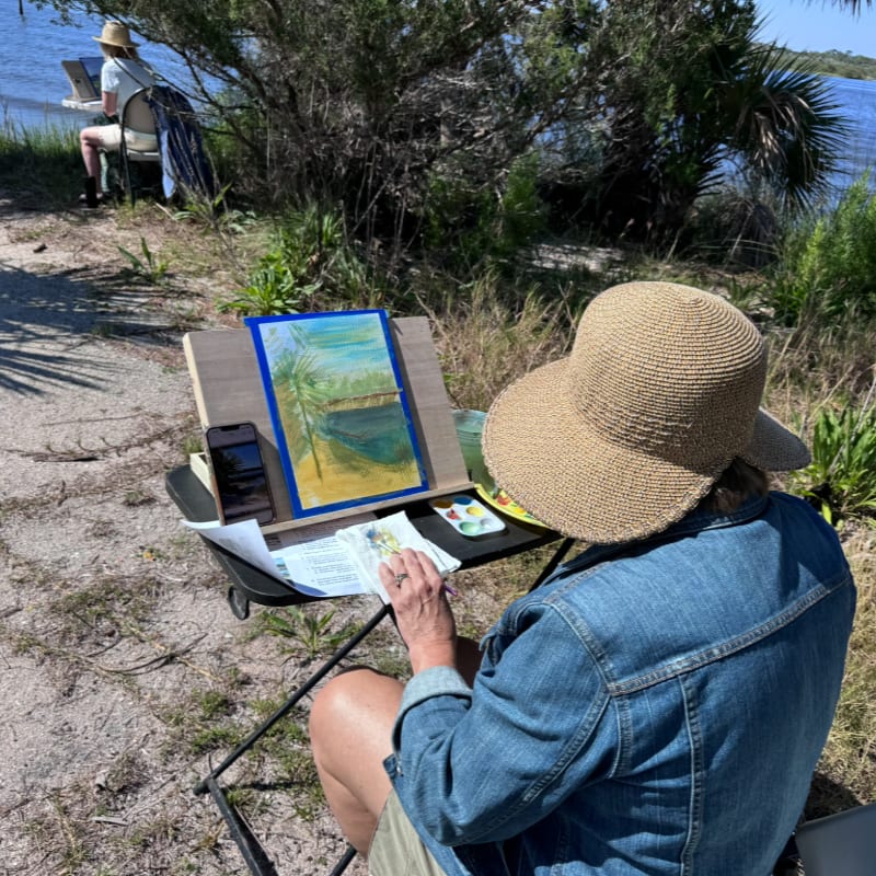 A person in a straw hat and denim jacket paints a landscape on an easel in the foreground, with another artist and a waterfront shoreline in the background.