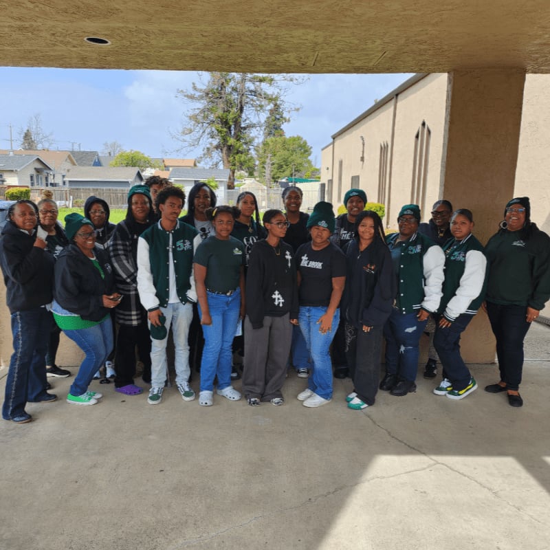 A group of people wearing green and black clothing stand together under a concrete overhang with a residential neighborhood and trees in the background.
