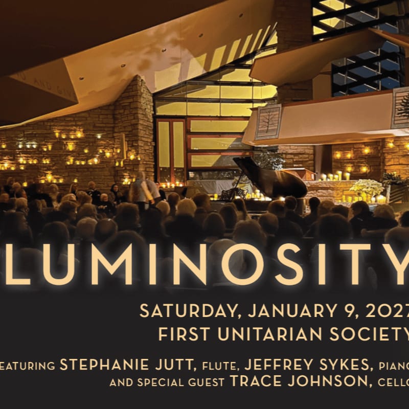 A crowd of people sits in a dimly lit, modern stone hall facing a grand piano and a brightly lit stage.
