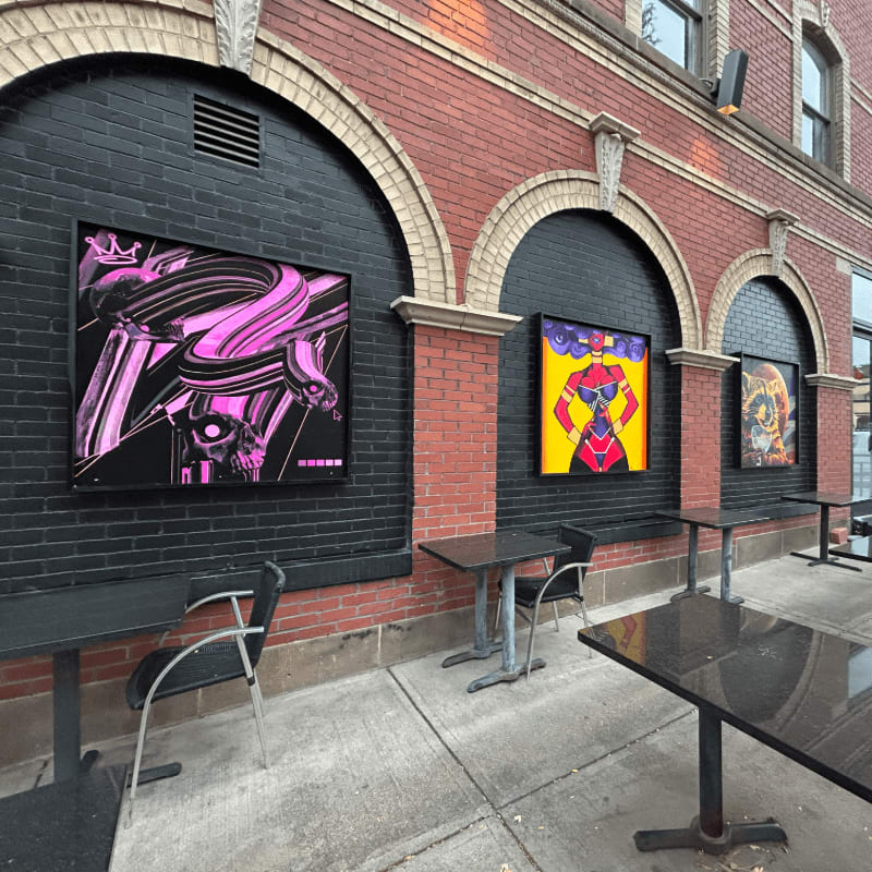 Three colorful art pieces hang on black walls within brick arches, with small tables and chairs set up on the sidewalk in the foreground.