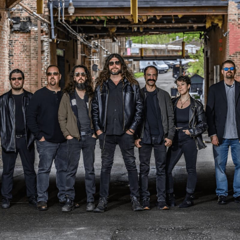 Seven people stand in a line in the foreground against the backdrop of a rustic, industrial alleyway.