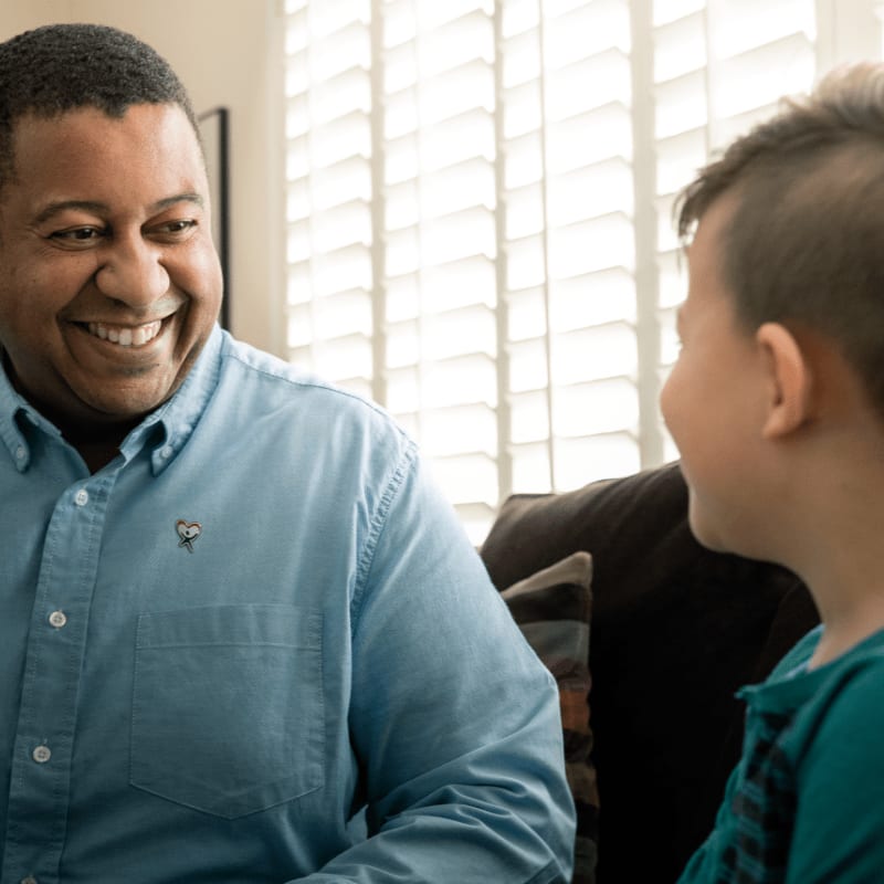 A smiling man in a blue shirt looks at a young boy in front of a window with white shutters.