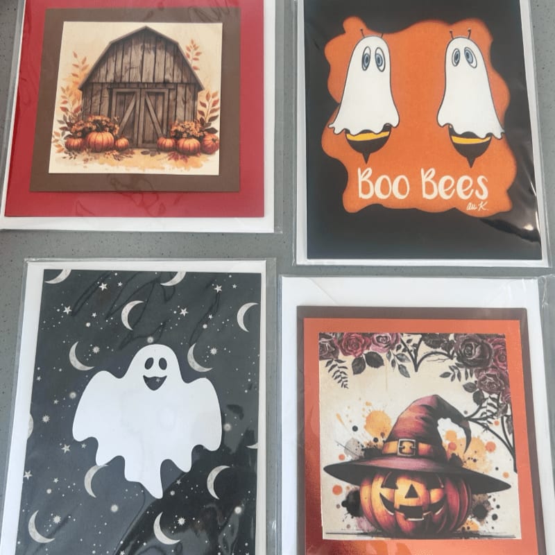 Four Halloween-themed greeting cards featuring a barn, ghost bees, a single ghost, and a pumpkin with a witch's hat are arranged on a white surface.