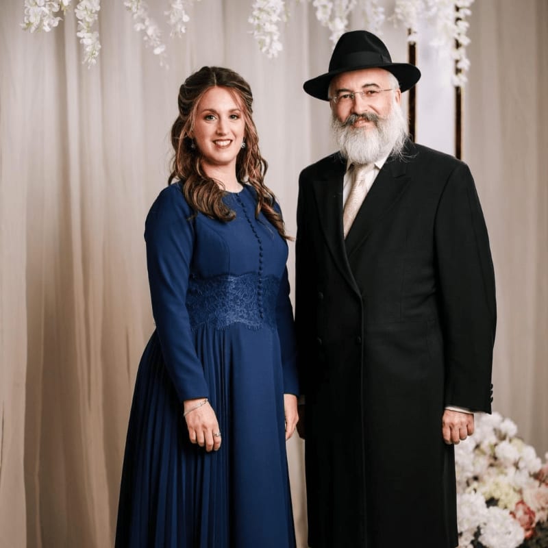 A woman in a long blue dress and a man in a black suit and hat stand together against a backdrop of neutral curtains and white floral decorations.