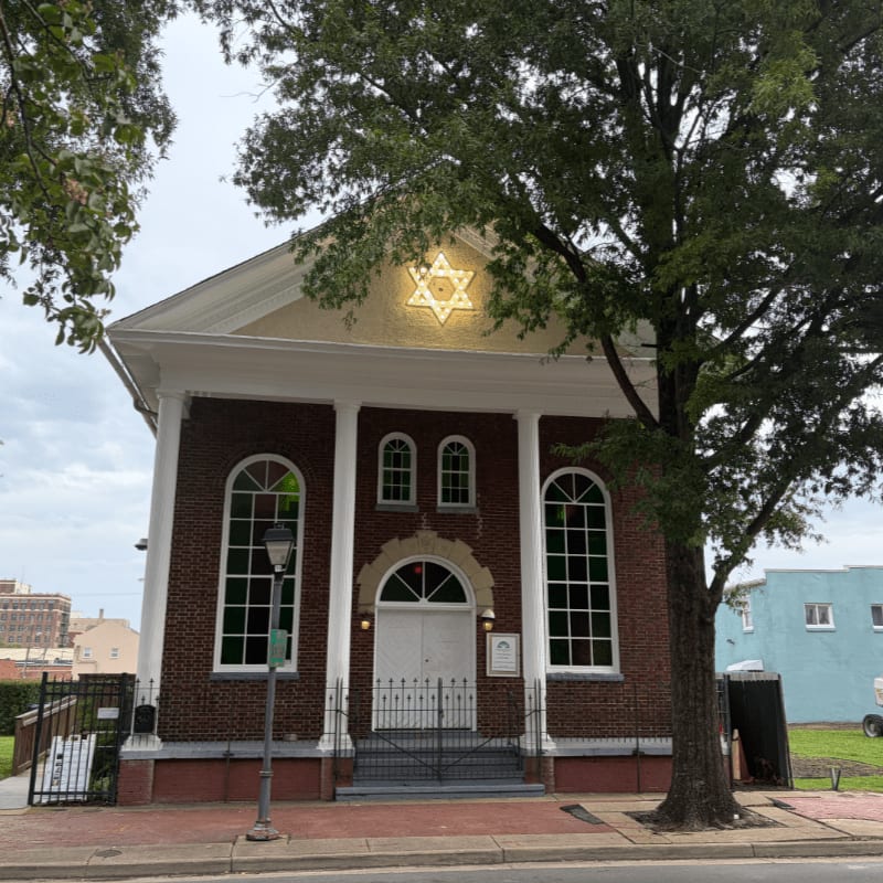 A brick synagogue with a Star of David on its pediment stands in the foreground, framed by large green trees against a cloudy sky.