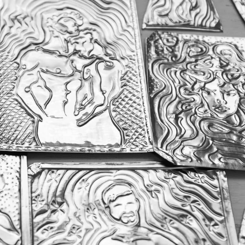 Embossed silver metal plaques featuring mythological figures are arranged in the foreground against a plain, dark background.