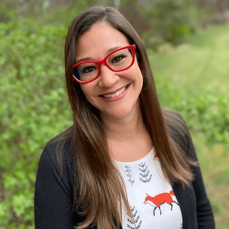 A smiling woman wearing red glasses and a fox-print shirt stands before a blurred background of green foliage.