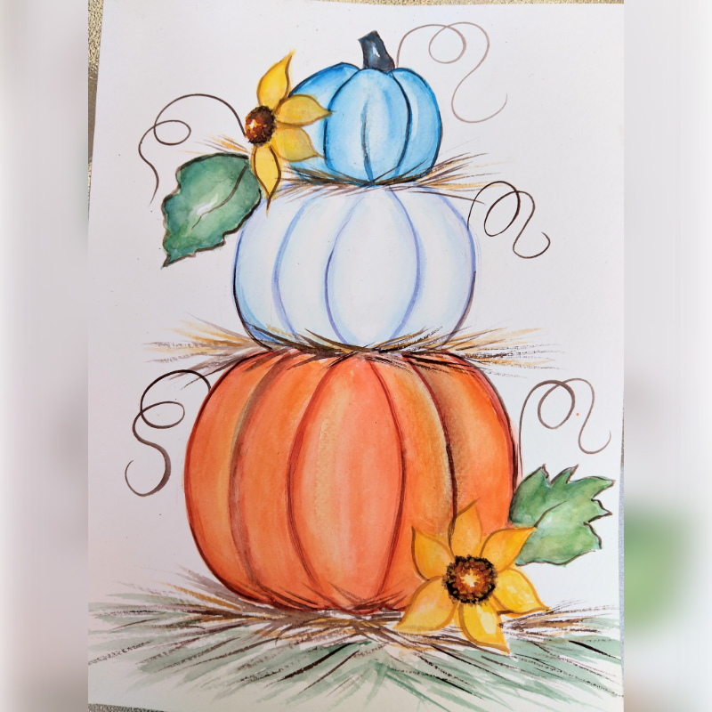 Three stacked pumpkins in orange, white, and blue are adorned with yellow sunflowers and green leaves against a plain white background.
