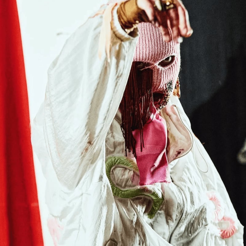 A person wearing a pink knit mask and a white oversized jacket stands against a background of a red curtain and a dark wall.