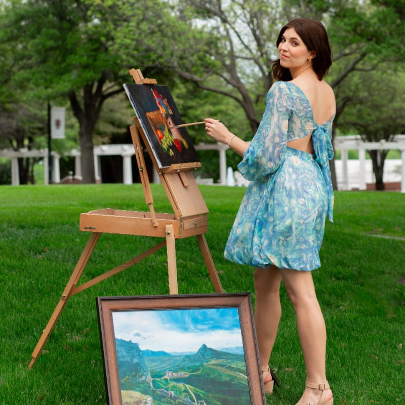 A woman in a blue patterned dress paints on an easel in the foreground, with a landscape painting resting on the grass and lush green trees in the background.