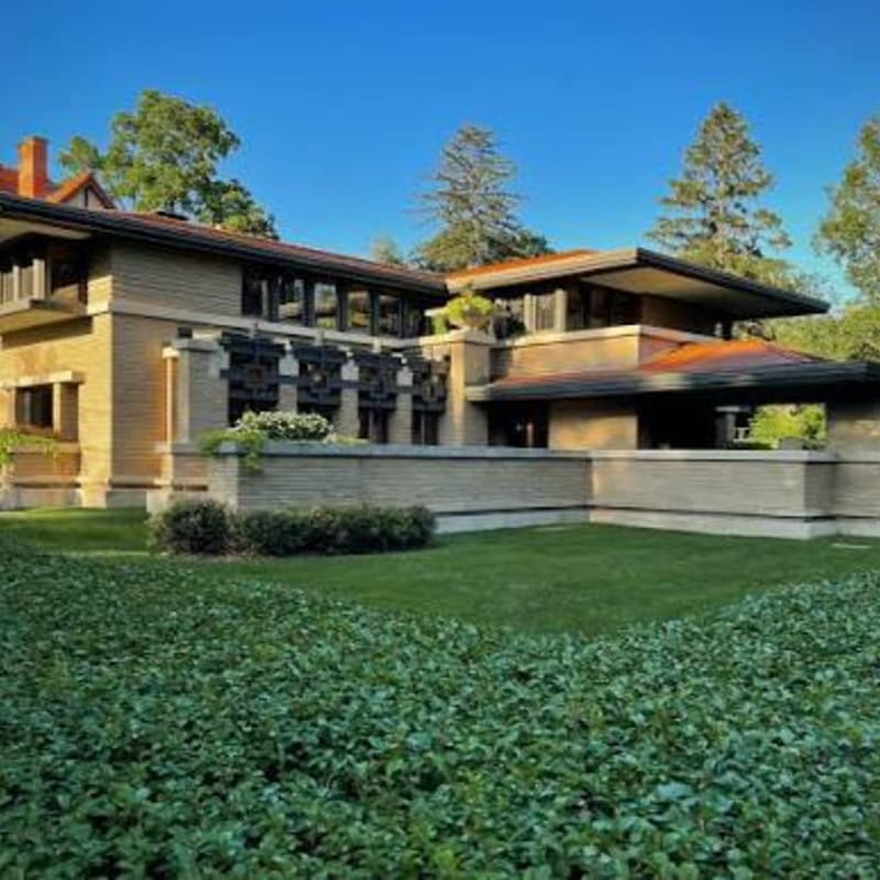 A modern prairie-style house sits behind a manicured green lawn and shrubbery under a clear blue sky with tall trees in the background.