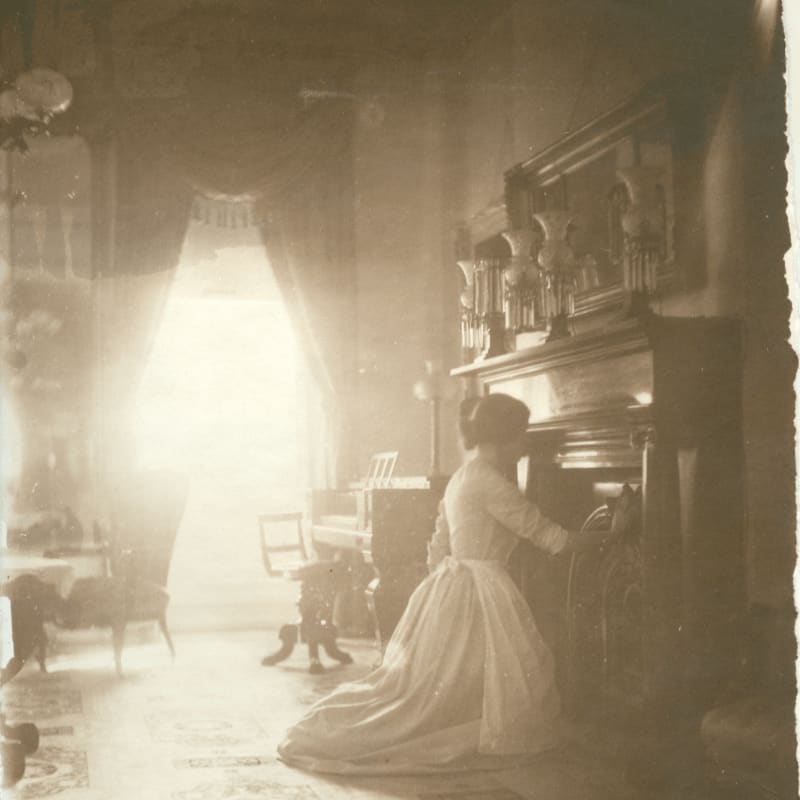 A woman in a long white dress kneels before a fireplace in the foreground, with a piano and a bright, curtained window in the background.
