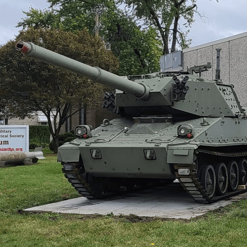A large, olive-drab tank with a long barrel sits on display in front of a building with trees and a sign.
