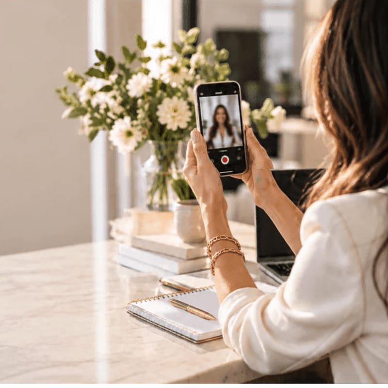 A woman in a white blouse holds a smartphone during a video call, with a laptop, notebook, and a vase of white flowers on a marble table in the background.