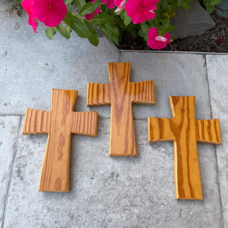 Three wooden crosses sit on a grey stone surface with pink flowers and green leaves in the background.