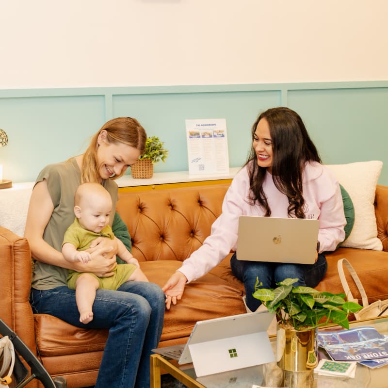Two women and a baby sit on a brown leather sofa in a room with light blue walls and a coffee table in the foreground.