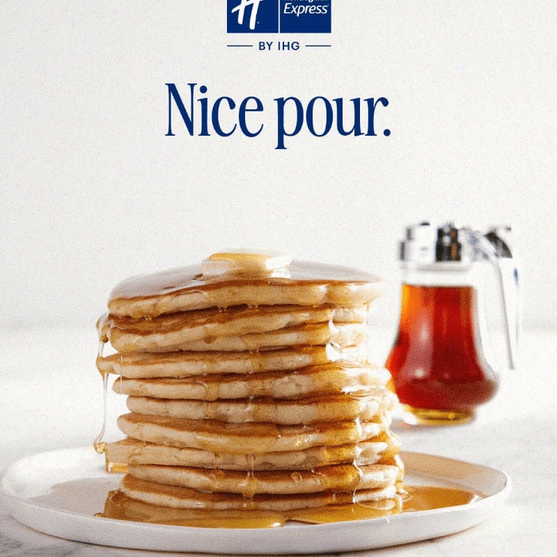 A tall stack of syrup-covered pancakes sits on a white plate in the foreground, with a syrup pitcher and a white background behind it.