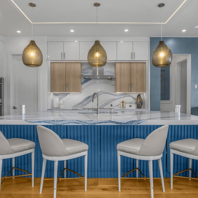 Four white bar stools sit before a blue ribbed kitchen island, with white and wood cabinetry and three glass pendant lights in the background.