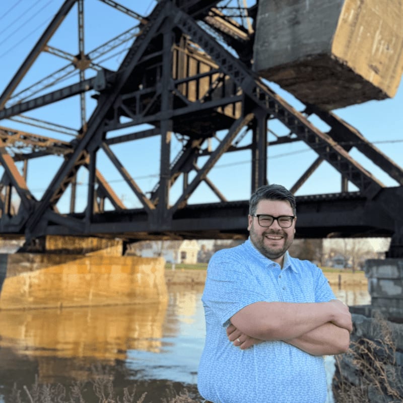 A man in a blue patterned shirt stands with his arms crossed in the foreground, while a large, rusty railway bridge spans the river in the background.