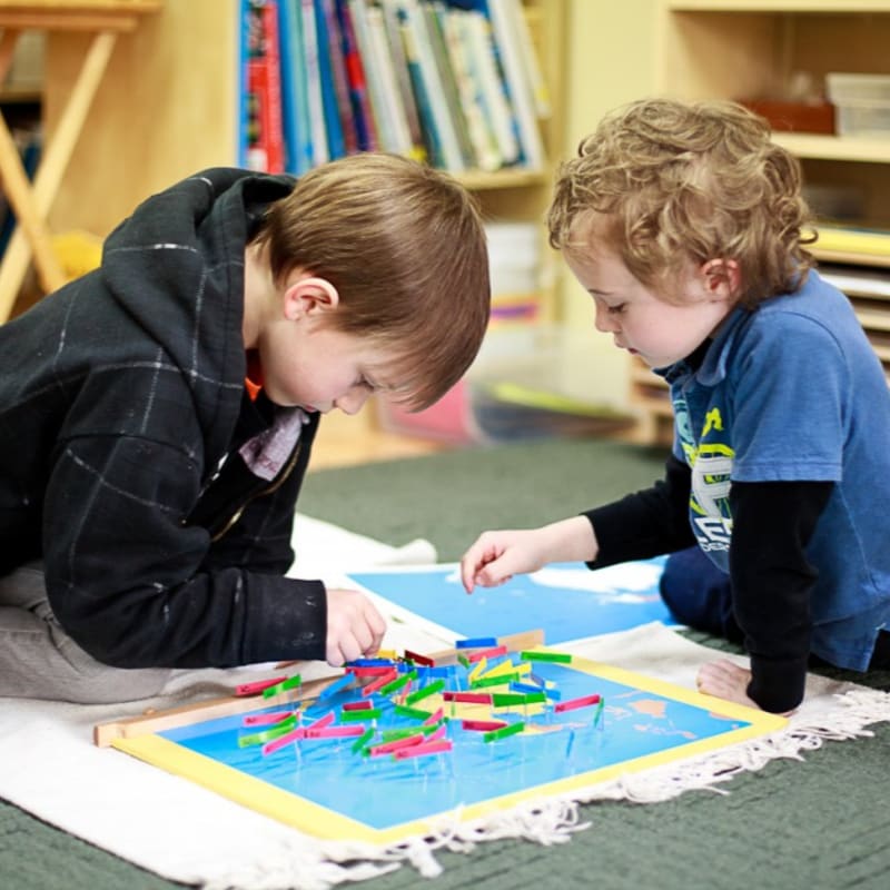 Two young boys play with colorful sticks on a blue board in the foreground, with bookshelves and a wooden table in the background.