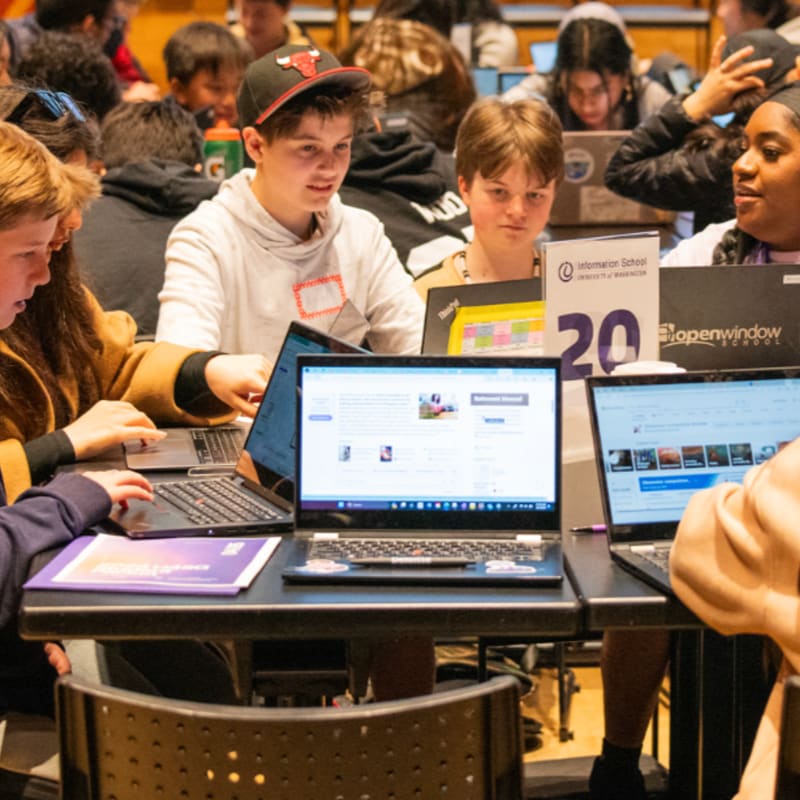 Students work on laptops at a table in the foreground, while a crowd of other students fills the background of a classroom.