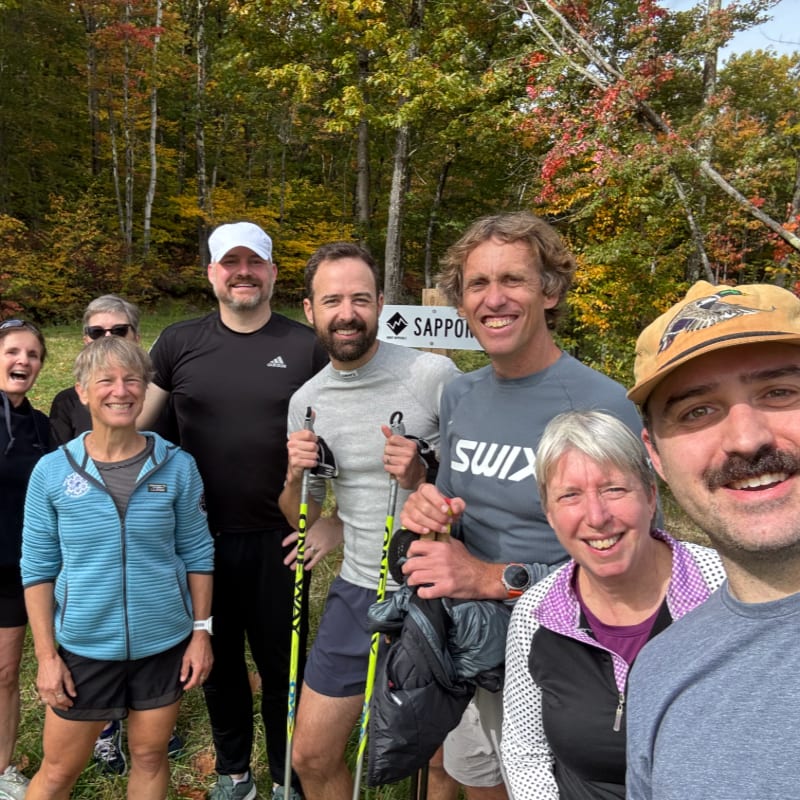 A group of smiling people pose for a photo in the foreground against a backdrop of autumn trees with yellow and red leaves.