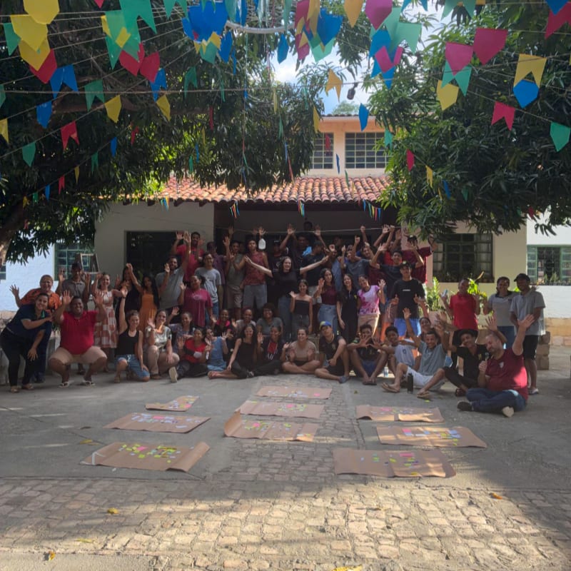 A large group of people pose together in a courtyard with colorful banners overhead and brown paper sheets spread across the ground in front of them.