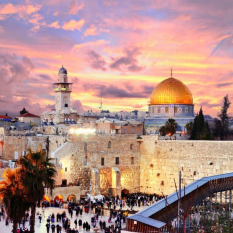 A crowd of people gathers before the Western Wall in the foreground, while the Dome of the Rock and a minaret stand against a vibrant sunset sky in the background.