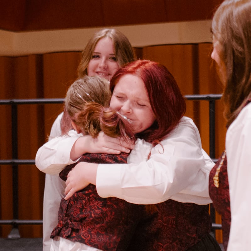 Two girls in white shirts and red patterned vests share an emotional hug in the foreground, while another smiling girl and a partial figure stand behind them against a brown paneled wall.