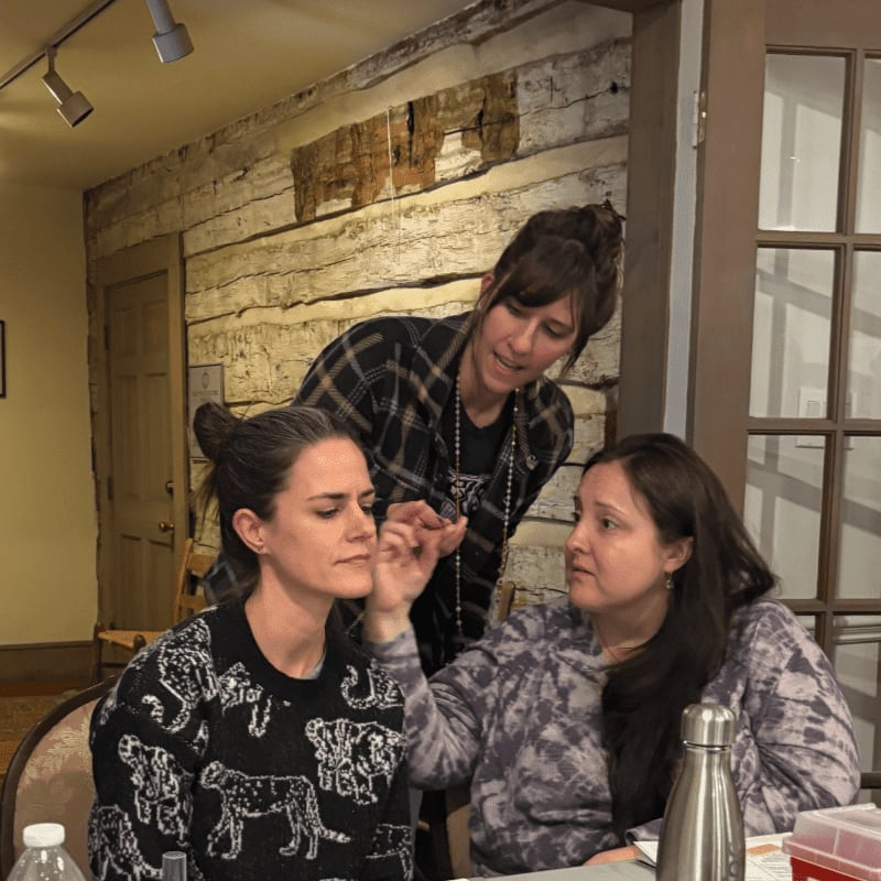 Three women are gathered around a table in a room with a rustic log wall and a glass-paned door in the background.