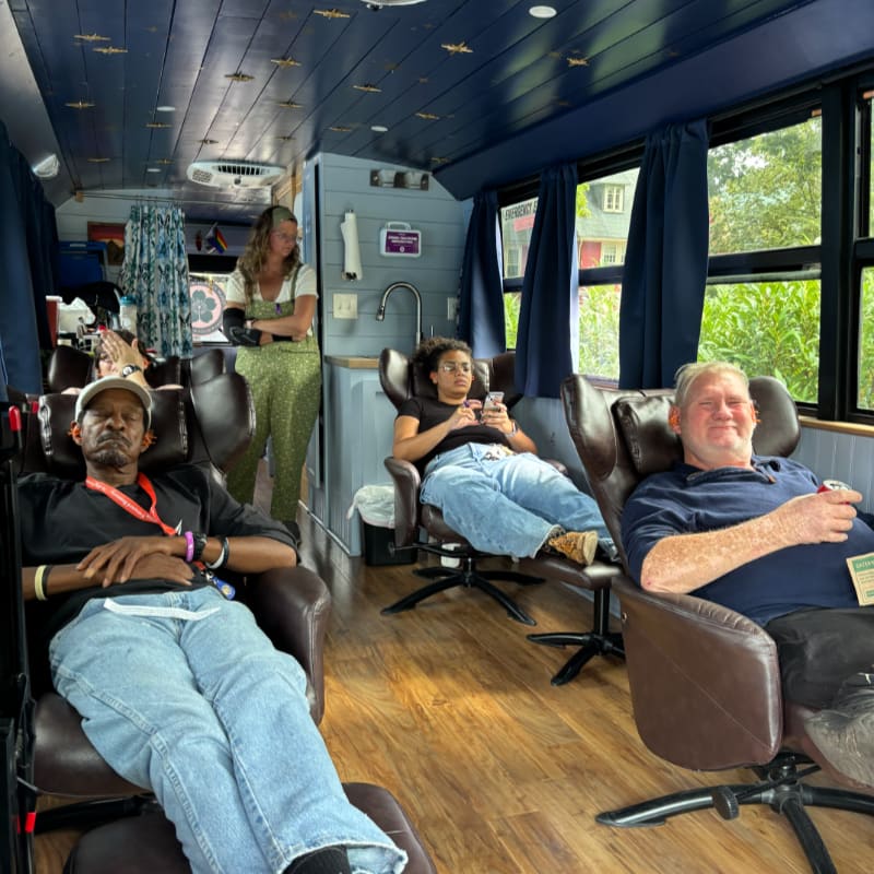 Four men relax in reclining chairs inside a bus with blue curtains and wooden floors, while a woman stands in the background near a sink and washing machine.