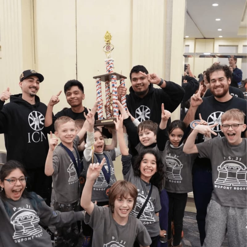 A group of smiling children and adults in matching black and grey shirts hold a tall trophy in a brightly lit indoor hallway.
