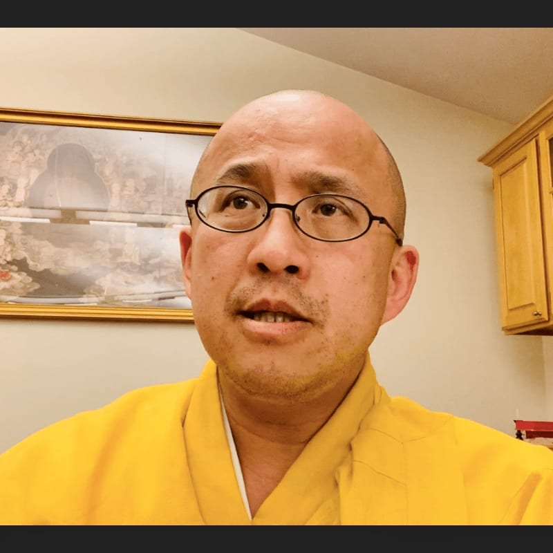 A bald man wearing glasses and a yellow robe stands in front of a framed picture and wooden cabinets.